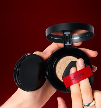 Close-up of a person's hands holding an open L'Oréal Paris cushion foundation compact. They are pressing a black sponge applicator with a red L'Oréal ribbon into the foundation-soaked cushion against a dark red background.
