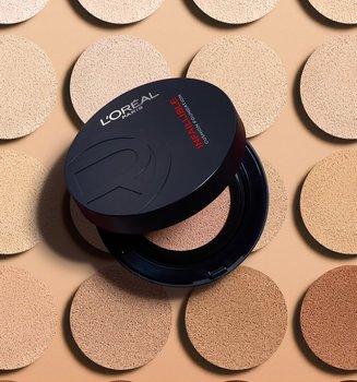 A sleek black L'Oréal Paris Infallible Cushion Foundation compact shown open, revealing its interior mirror and a black sponge applicator with a signature red ribbon. The product is displayed against a dark red gradient background.