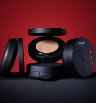 A collection of four L'Oréal Paris Infallible Cushion Foundation compacts arranged on a black surface against a bold red background. One compact in the center is open, revealing the foundation-soaked cushion, while others are closed, showcasing the sleek black packaging and product name.