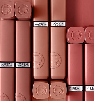 Multiple L'Oréal matte lipstick tubes in various nude and rose shades, arranged vertically side-by-side against a solid dark mauve background.