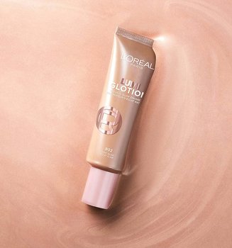 A tube of L'Oréal Lumi Glotion natural glow enhancer centered on a shimmery, iridescent rose-gold liquid background.