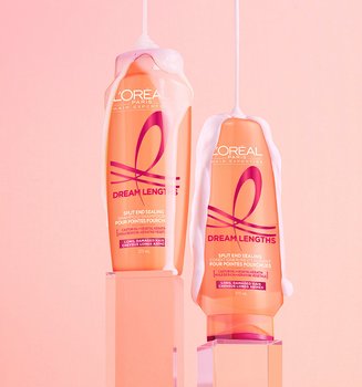 L'Oréal Paris Dream Lengths Shampoo and Conditioner bottles in peach-colored packaging, sitting on translucent pink pedestals with white foam dripping down against a soft pink background.