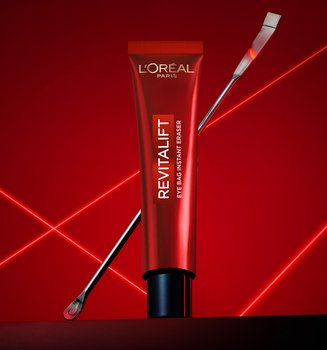 A professional product photography of the L'Oréal Paris Revitalift Eye Bag Instant Eraser. A sleek red tube stands upright in the center on a dark, reflective surface, with a thin metallic laboratory spatula leaning diagonally behind it. The composition is set against a deep red background illuminated by vibrant, glowing red laser lines that crisscross in an 'X' pattern, creating a high-tech, clinical, and scientific atmosphere that emphasizes the precision and dermatological expertise of the skincare product