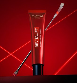 A professional product photography of the L'Oréal Paris Revitalift Eye Bag Instant Eraser. A sleek red tube stands upright in the center on a dark, reflective surface, with a thin metallic laboratory spatula leaning diagonally behind it. The composition is set against a deep red background illuminated by vibrant, glowing red laser lines that crisscross in an 'X' pattern, creating a high-tech, clinical, and scientific atmosphere that emphasizes the precision and dermatological expertise of the skincare product