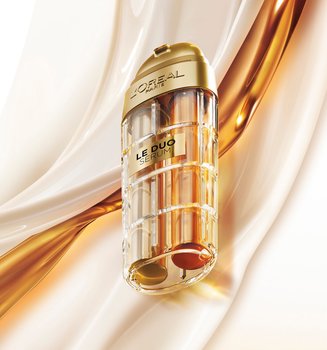 Commercial image of the L'Oréal Paris Le Duo Serum. The luxurious bottle, with gold accents and a transparent body revealing two distinct formulas (one clear and one amber), is presented at a diagonal angle. The background consists of smooth, silky waves of white and golden textures swirling together, symbolizing the power and richness of the serum blend.
